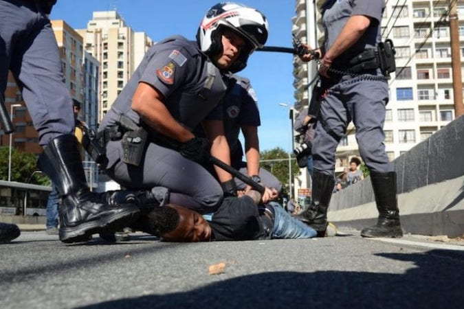 Police officer with a club forcibly restraining a Black man who lies face down on the pavement while two other officers observe.