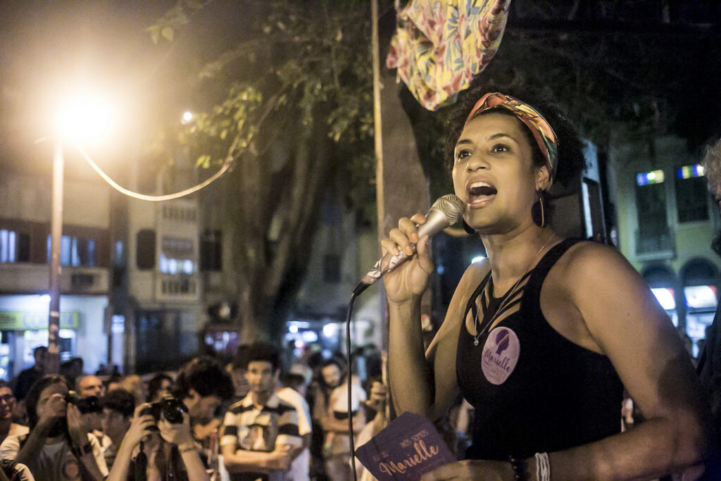 A Black woman speaks into a microphone in front of a crowd gathered outside at night. A sticker on her shirt and pamphlet in her hand read "Marielle."