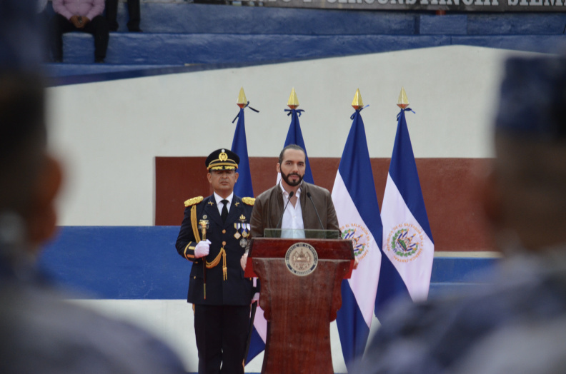 Seen from the audience, a man speaks from an official podium with a uniformed officer and four El Salvadorian flags behind him.