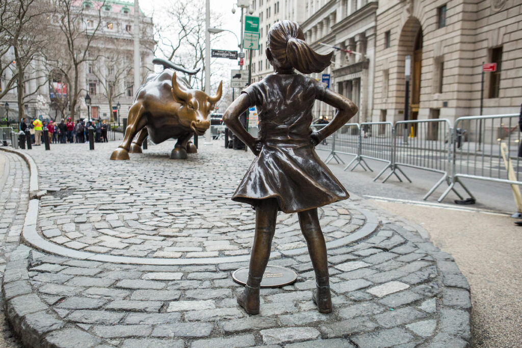 Statue of young girl in a skirt, legs astride and hands on hips, faces down a statue of a charging bull opposite.