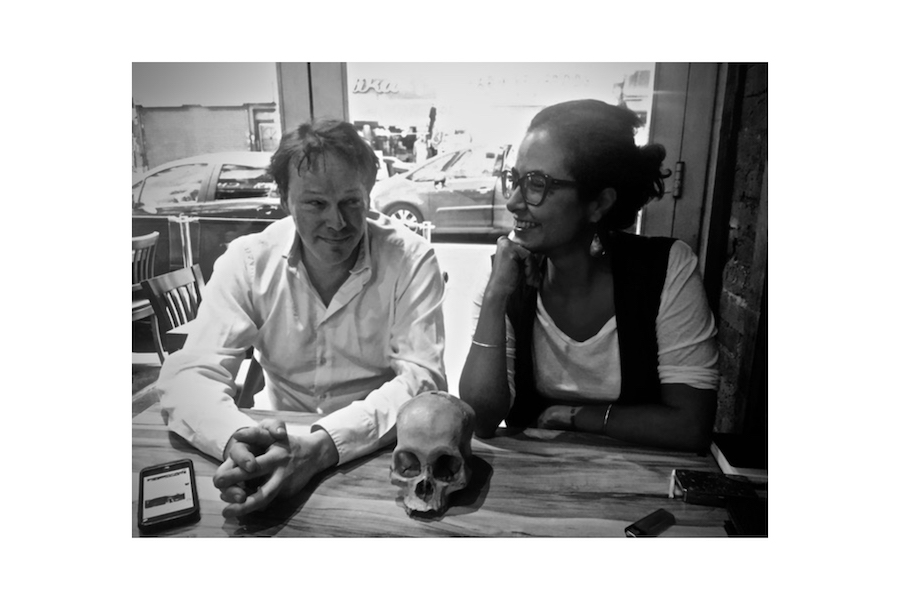 Black and white photo of a man and woman sitting together at a table, smiling, with a skull on the table between them.