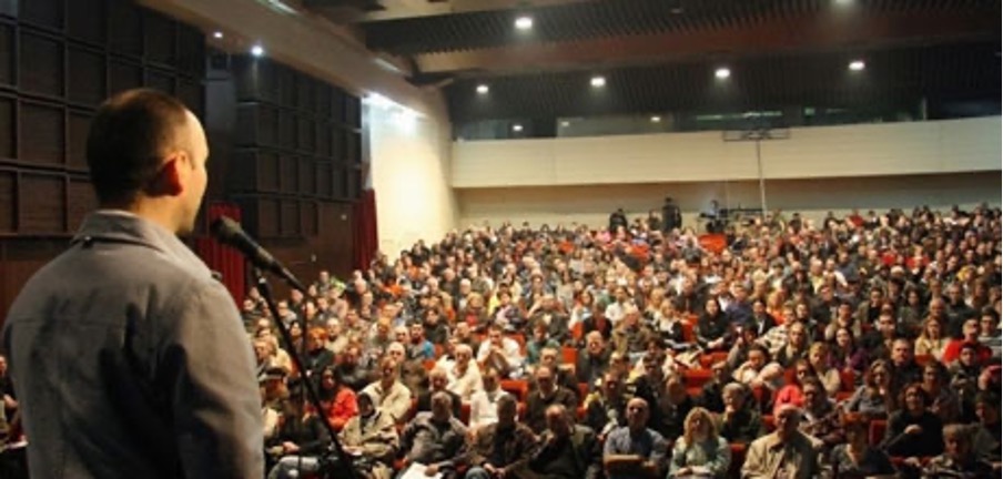 A man at a microphone speaking to a crowded room.