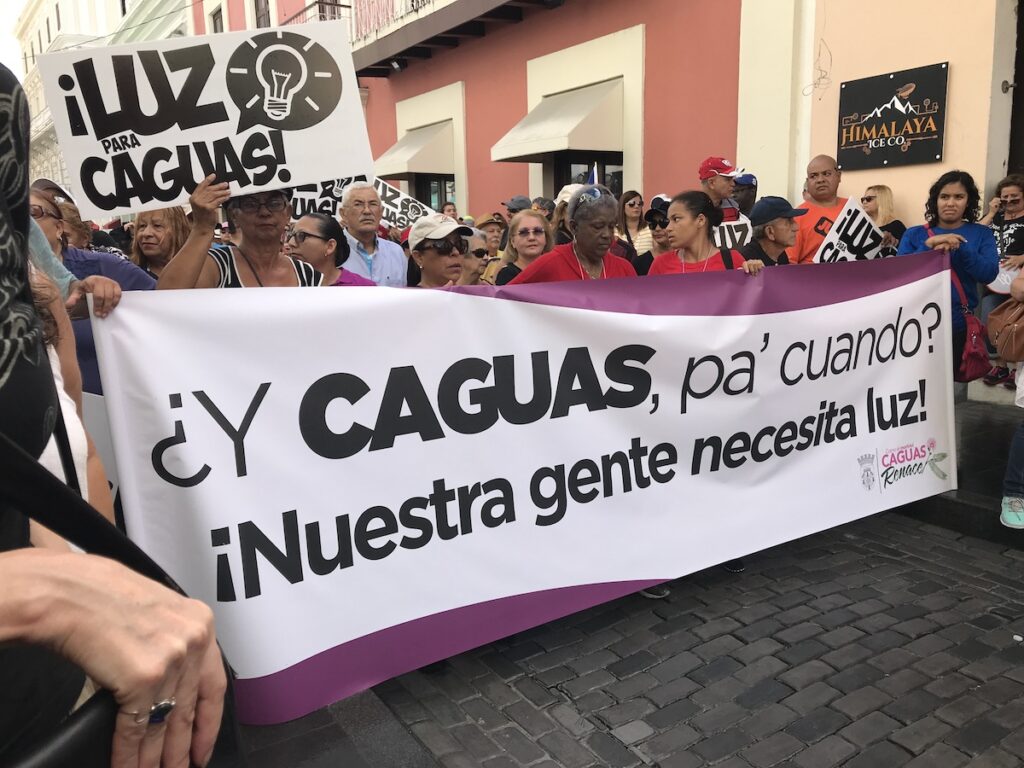 Protesters march down a street, holding signs that say "Luz para Caguas" and "¿Y Caguas pa' cuando? ¡Nuestra gente necesita luz!"