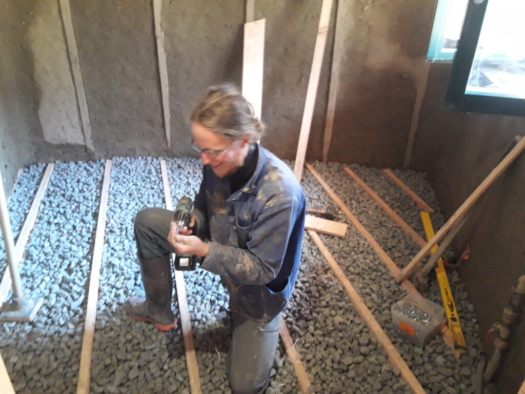 A blonde, white person in a dirty jacket and work boots kneels on a gravel floor lined with boards, holding a power drill.