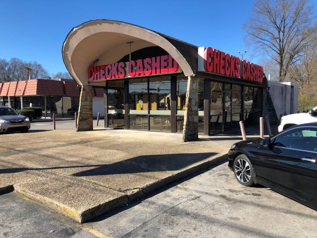 A large sign reads "CHECKS CASHED" on a small building with a curved roof, stone facade, and glass walls in a parking lot.