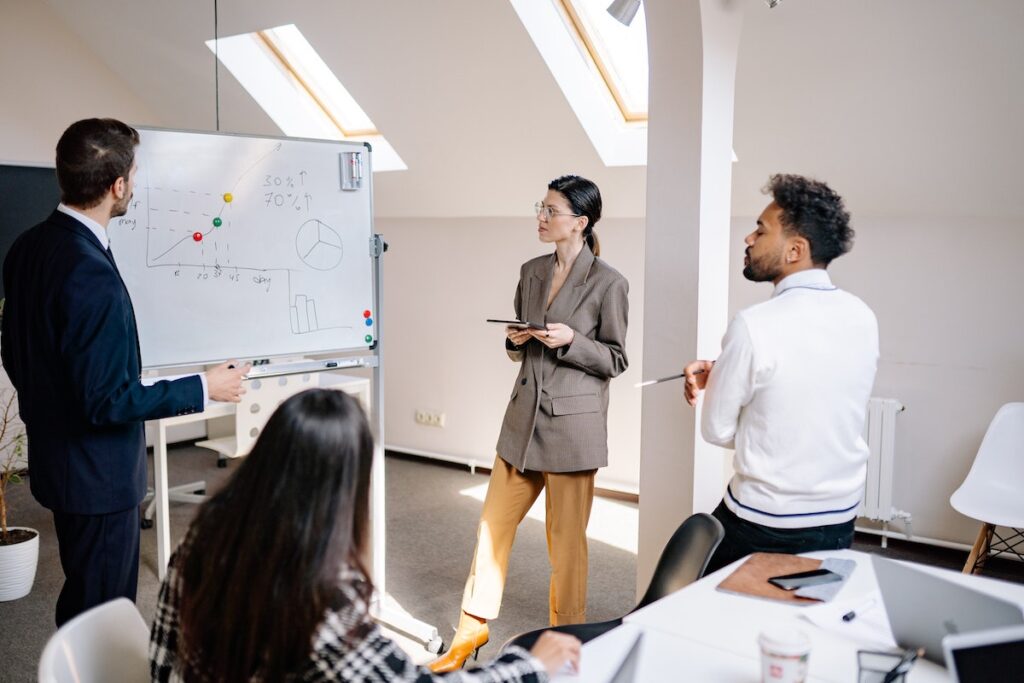 Two men and two women, dressed in business attire, look at a whiteboard with a graph showing an upward trend.