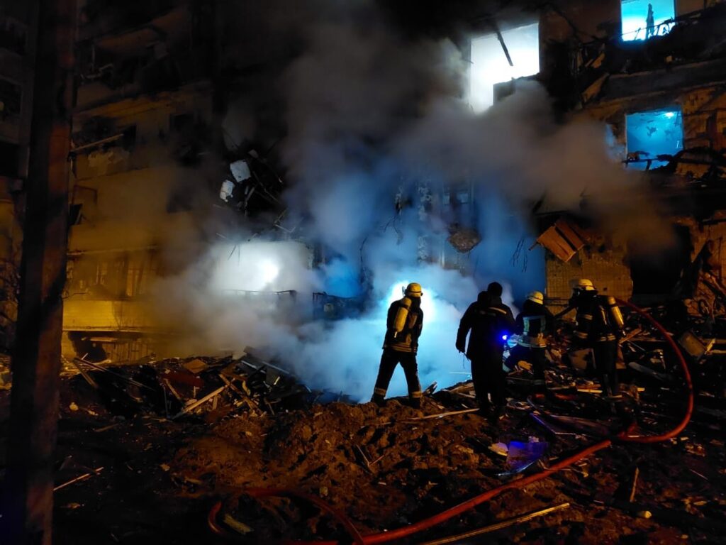 Dimly lit firefighters stand amid smoke and ruined buildings.