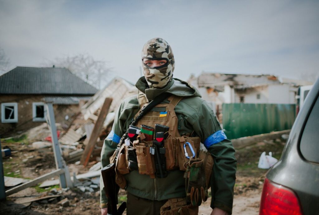 A white person stands in front of a destroyed house, wearing a camo balaclava, green jacket, and ammo and gear vest with a gun strapped across one shoulder. A Ukrainian flag is displayed on the chest of the gear vest.