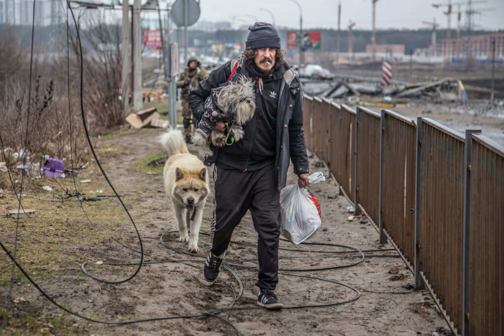 A white man wearing a gray beanie and black tracksuit, with a cigarette in his mouth, carries a small dog and a grocery sack down a pathway surrounded by trash and rubble. A larger dog walks behind him and others follow in the distance.